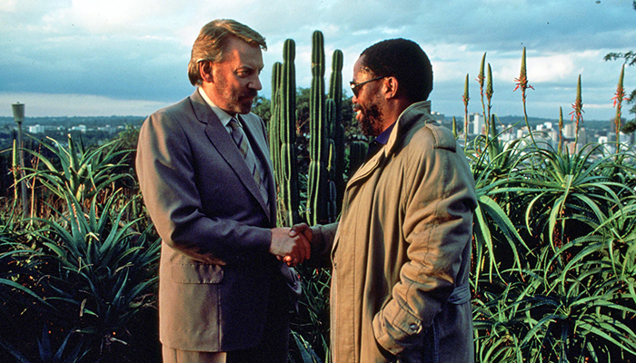 A white man and a Black man stand shaking hands in front of a large cactus.