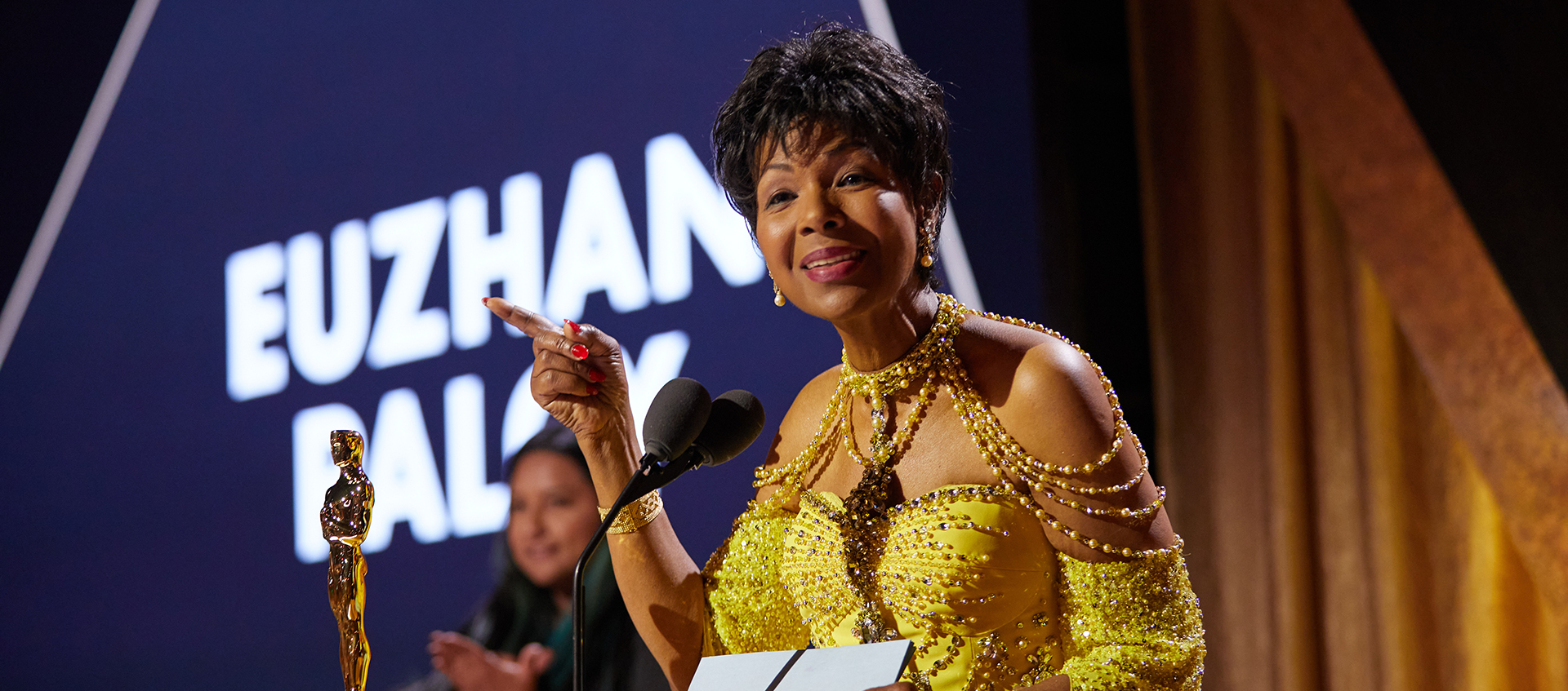 A Black woman wearing a formal dress stands at a podium with an Oscar statue on it.