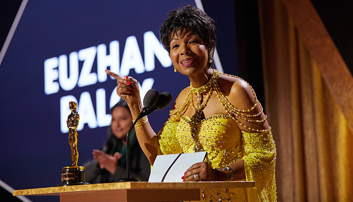 A Black woman wearing a formal dress stands at a podium with an Oscar statue on it.