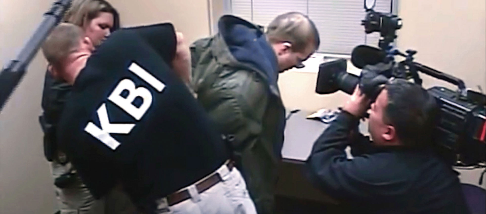 A man with the acronym KBI on his shirt arrests another man. A third man records the arrest.