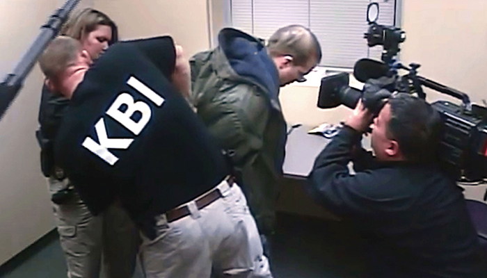 A man with the acronym KBI on his shirt arrests another man. A third man records the arrest.