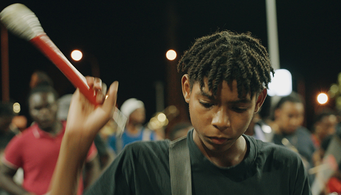 A young Black boy looks downward and raises a drumstick. A group of people are behind him.