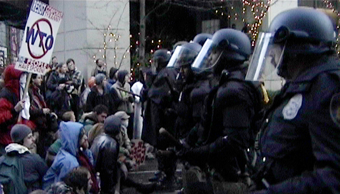 Police in riot gear face off with protesters in the street.