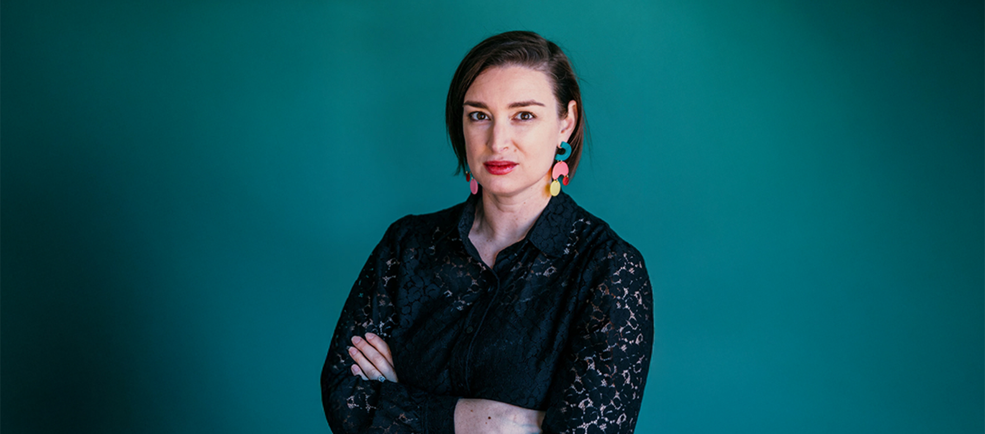 A headshot of Leah Triplett, who has light skin and short brown hair. She wears a black blouse and colorful earrings.