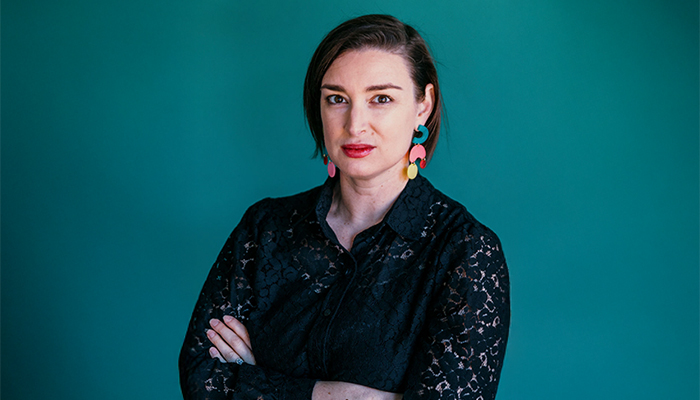A headshot of Leah Triplett, who has light skin and short brown hair. She wears a black blouse and colorful earrings.