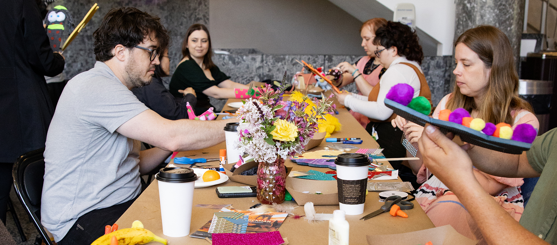 People sit at a long table filled with art and craft supplies. Each person works on creating their own artwork.