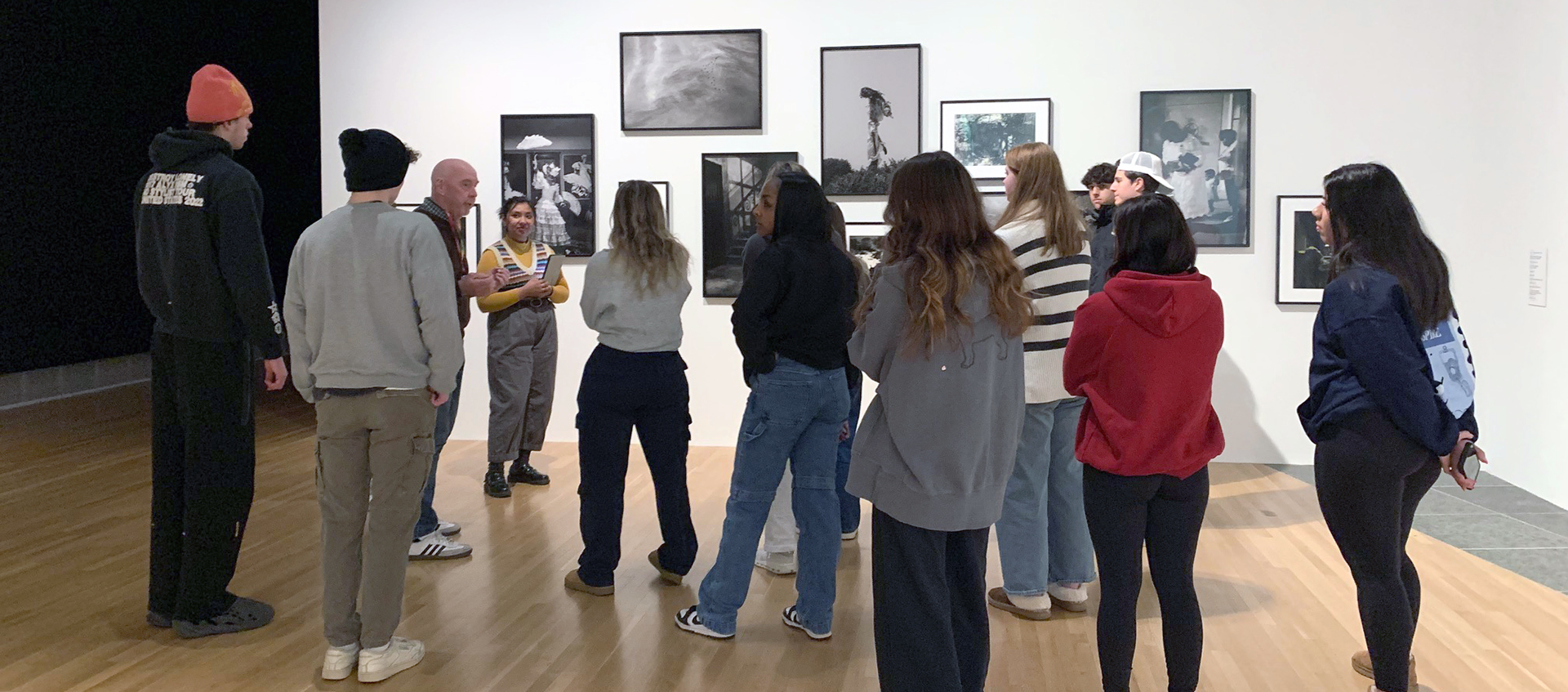A group of students stand in a gallery space looking at framed photographs hung on a white gallery wall.