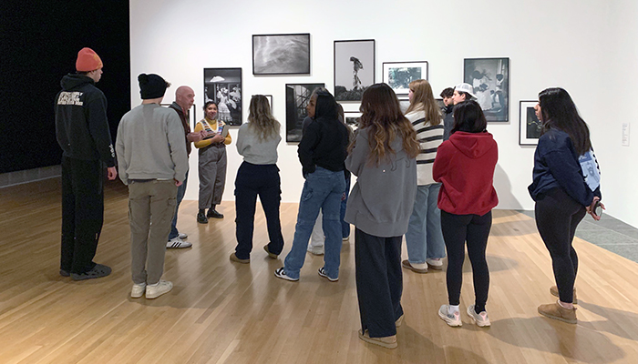 A group of students stand in a gallery space looking at framed photographs hung on a white gallery wall.