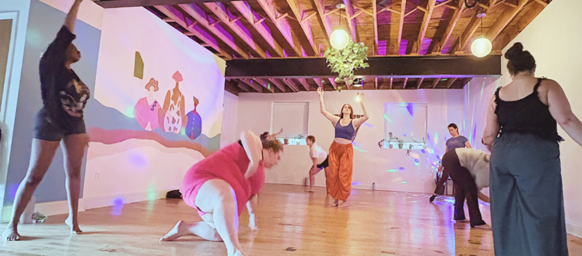 People move expressively in an open gallery space lit by overhead  lights, a spinning rainbow-colored globe table lamp, and natural light. 