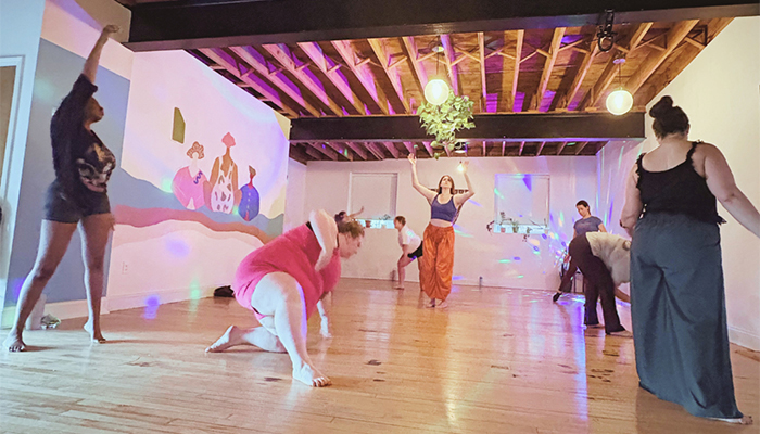 People move expressively in an open gallery space lit by overhead  lights, a spinning rainbow-colored globe table lamp, and natural light. 