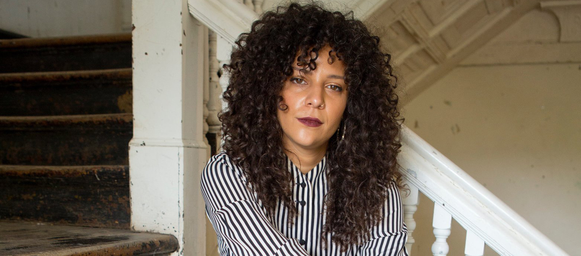Sofía Córdova sits on a worn wooden staircase. She has light brown skin, long, curly, dark-brown hair, and wars a striped button-down shirt.