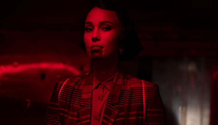 A woman wearing a plaid blazer, with a tattoo on her chin in a red-lit room.