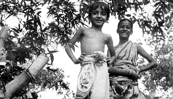 Two children in traditional clothing stand outdoors among trees, smiling and looking at the camera.