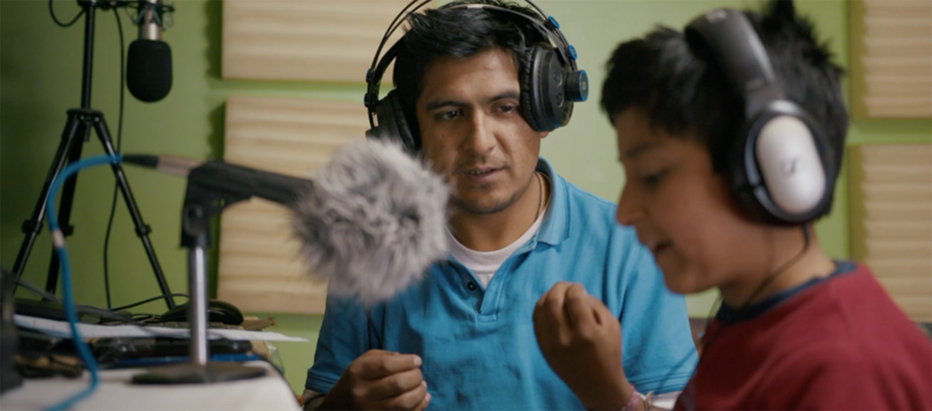 A man and a boy both wearing headphones sit at a desk with microphones.
