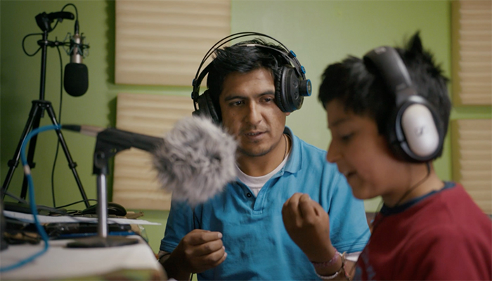 A man and a boy both wearing headphones sit at a desk with microphones.
