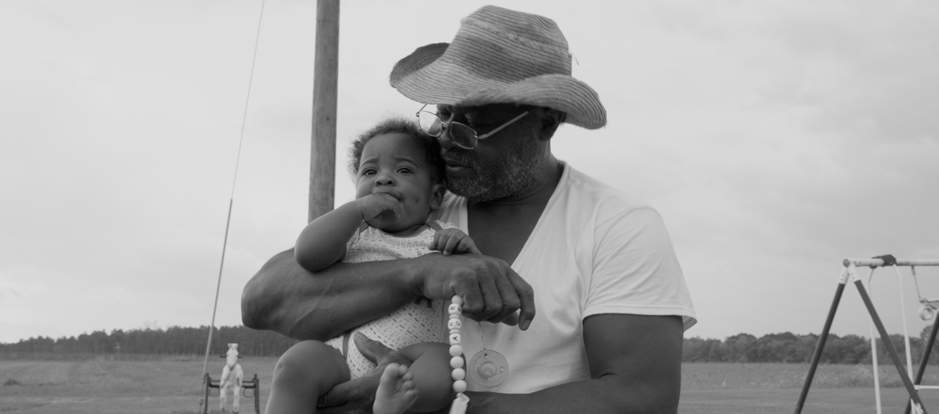 An older man wearing a hat and glasses holds a baby outdoors in a grassy field, with playground equipment visible in the background.