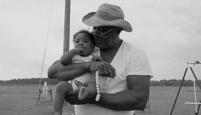An older man wearing a hat and glasses holds a baby outdoors in a grassy field, with playground equipment visible in the background.