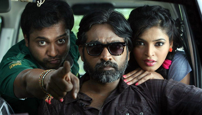 Three people sit closely together in a car; the man in the center wears sunglasses, the man on the left points forward, and the woman on the right leans on the central man's shoulder.