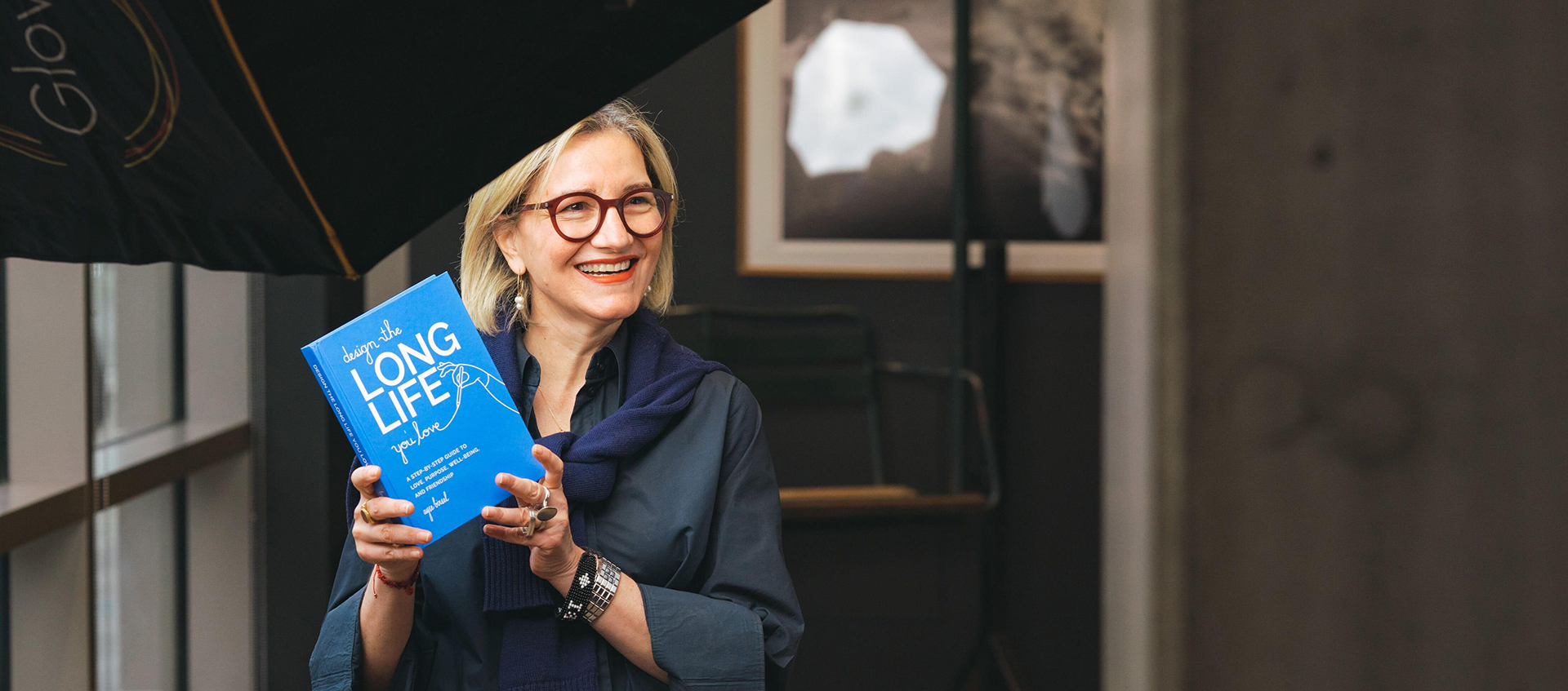 Ayse Birsel smiles while holding her book titled Design the Long Life You Love.