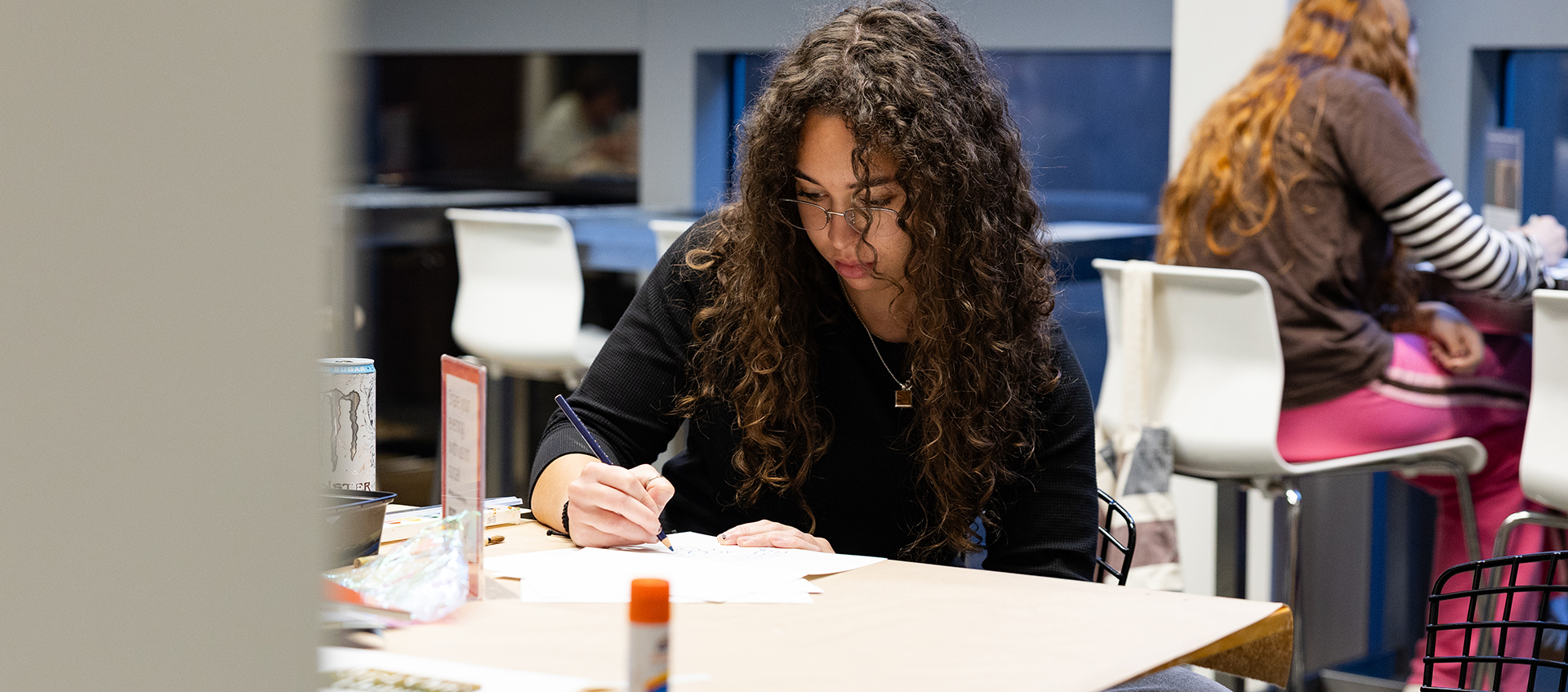 A young woman with long curly brown hair sits at a café table and draws on a piece of paper.