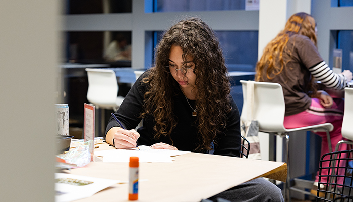 A young woman with long curly brown hair sits at a café table and draws on a piece of paper.