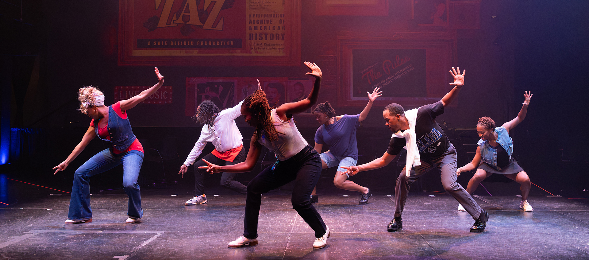 Six dancers perform a synchronized routine on stage, each striking a dynamic pose with one arm raised and one bent, under colorful stage lighting.