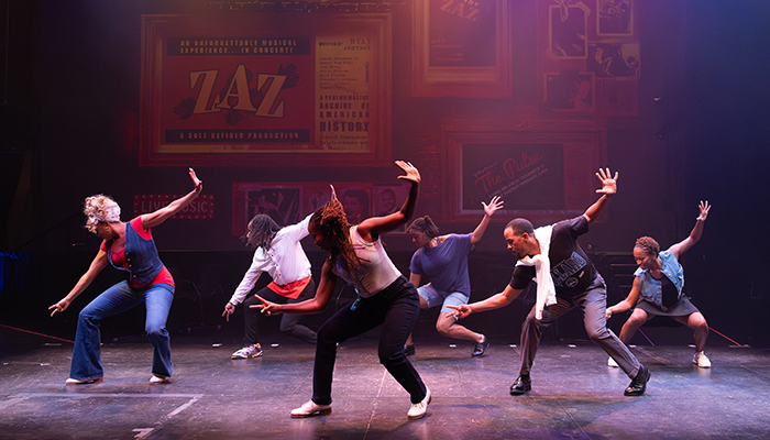 Six dancers perform a synchronized routine on stage, each striking a dynamic pose with one arm raised and one bent, under colorful stage lighting.