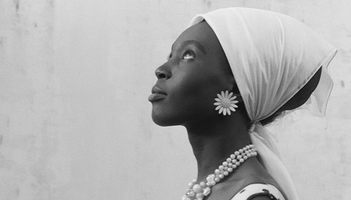A black-and-white still of the profile of a young Black woman, who wears a head scarf, flower earrings, and a pearl necklace.