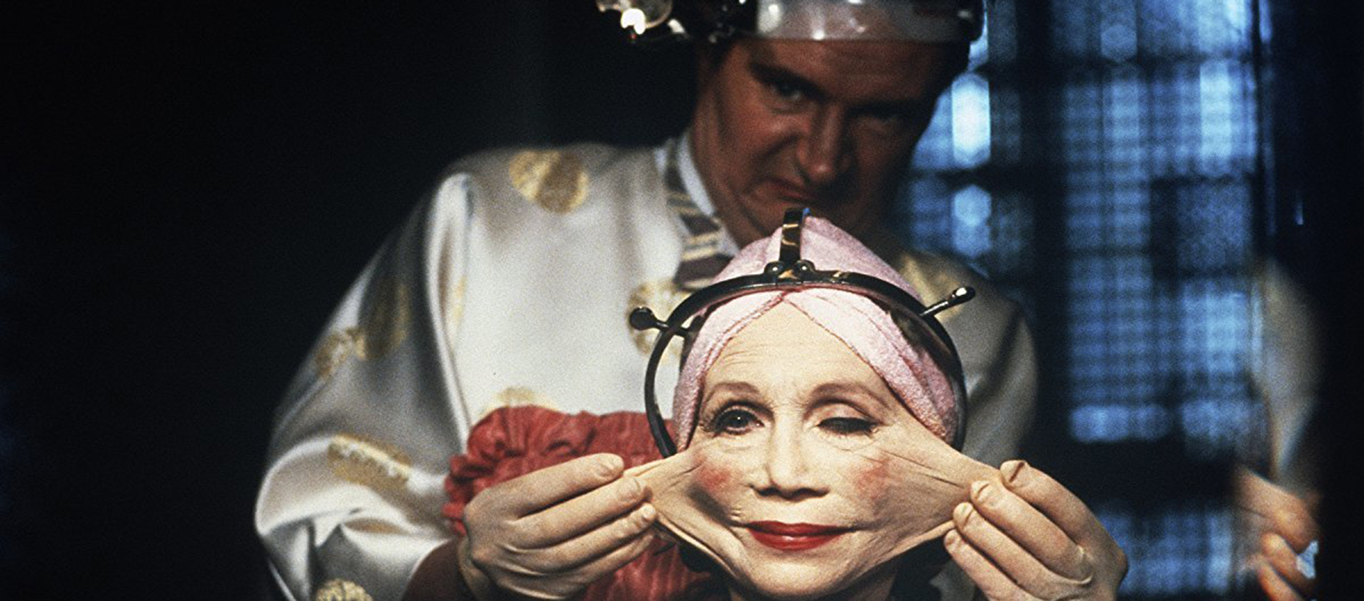 A man stretches an older person's cheeks widely. The older person wears makeup and a metal helmet.