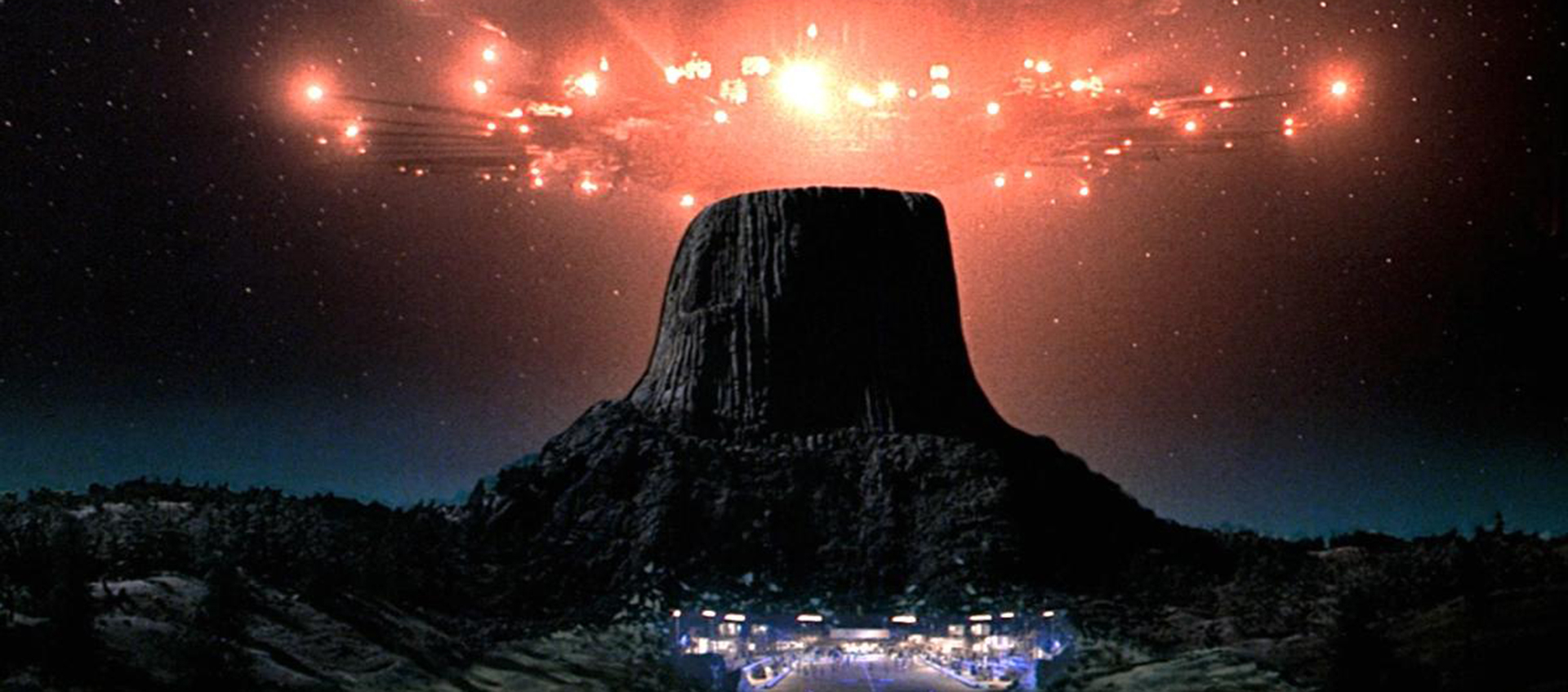 A brightly lit spaceship hovers over a mountain against a dark, story sky. A city is visible at the bottom of the mountain.