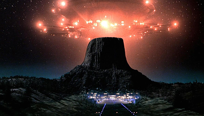 A brightly lit spaceship hovers over a mountain against a dark, story sky. A city is visible at the bottom of the mountain.