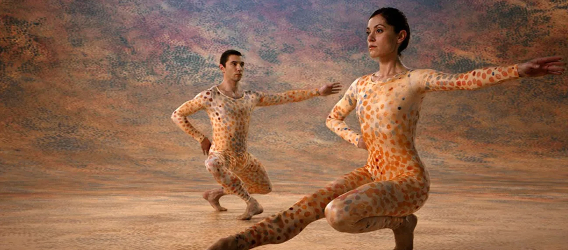Two dancers in colorful, spotted leotards squat in dance positions. They are surrounded by a colorful, spotted background.