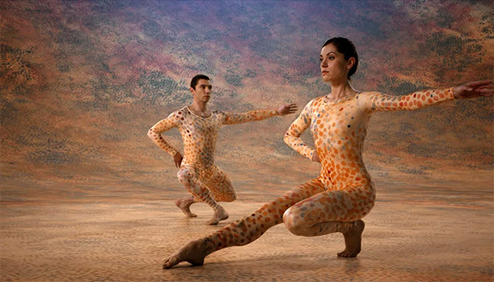 Two dancers in colorful, spotted leotards squat in dance positions. They are surrounded by a colorful, spotted background.
