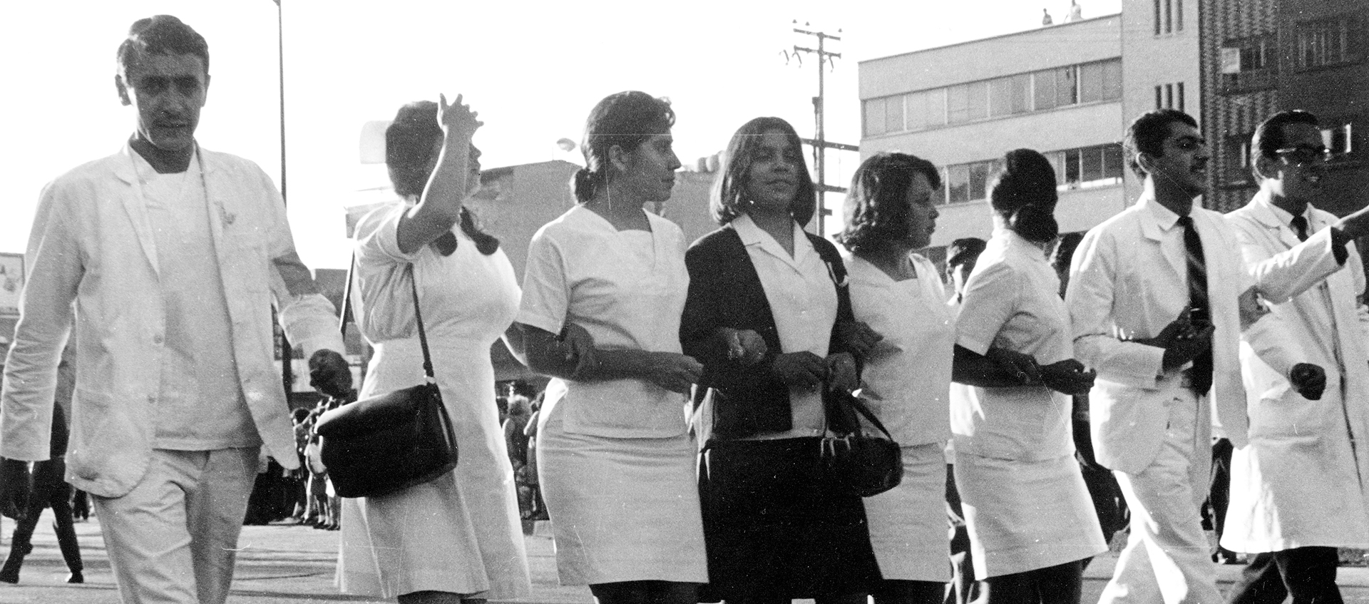 Black-and-white still of a row of people walking down a street. They all wear white.