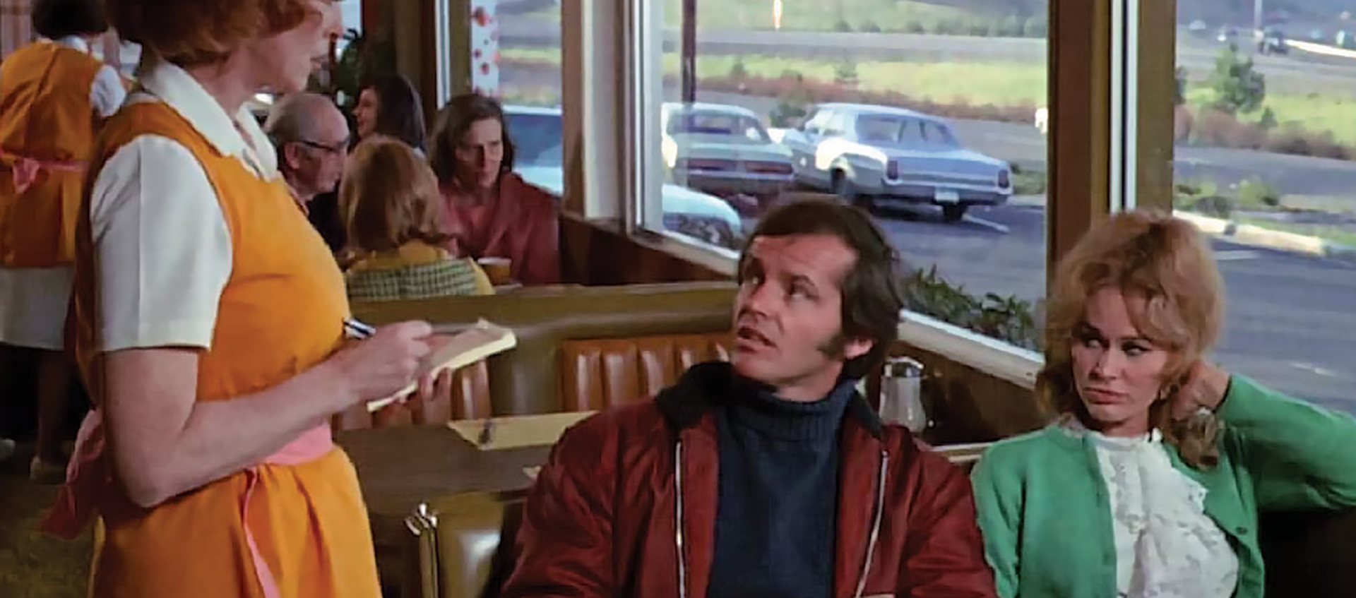 A white man and a white woman sit in a booth at a diner. A server takes their order.