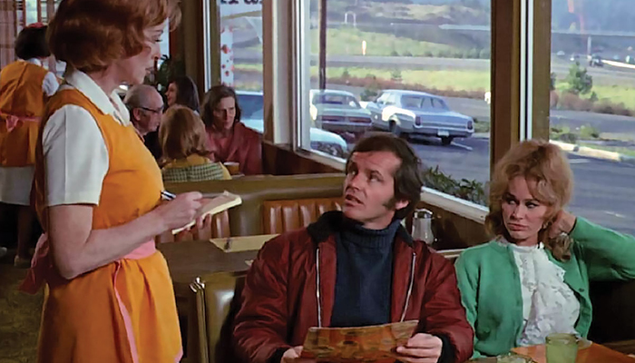 A white man and a white woman sit in a booth at a diner. A server takes their order.