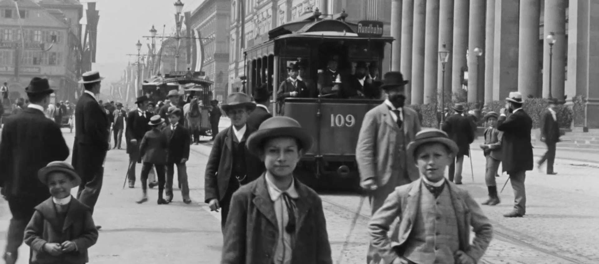 Black-and-white still of people on a busy street. There are children in the forefront looking at the camera.