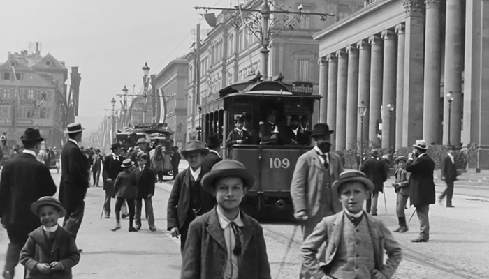 Black-and-white still of people on a busy street. There are children in the forefront looking at the camera.