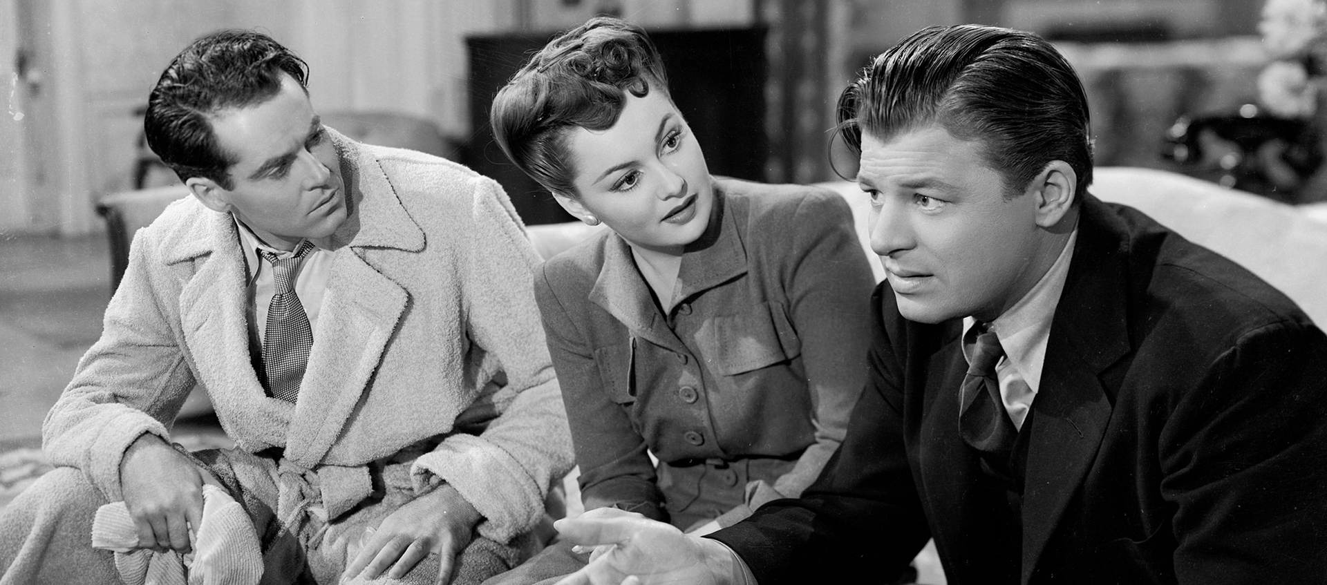 A black-and-white still of two white men and a white woman sitting on a couch and conversing. The men appear worried.