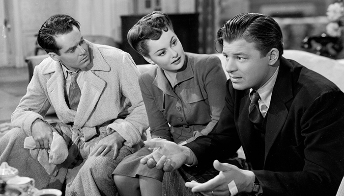 A black-and-white still of two white men and a white woman sitting on a couch and conversing. The men appear worried.