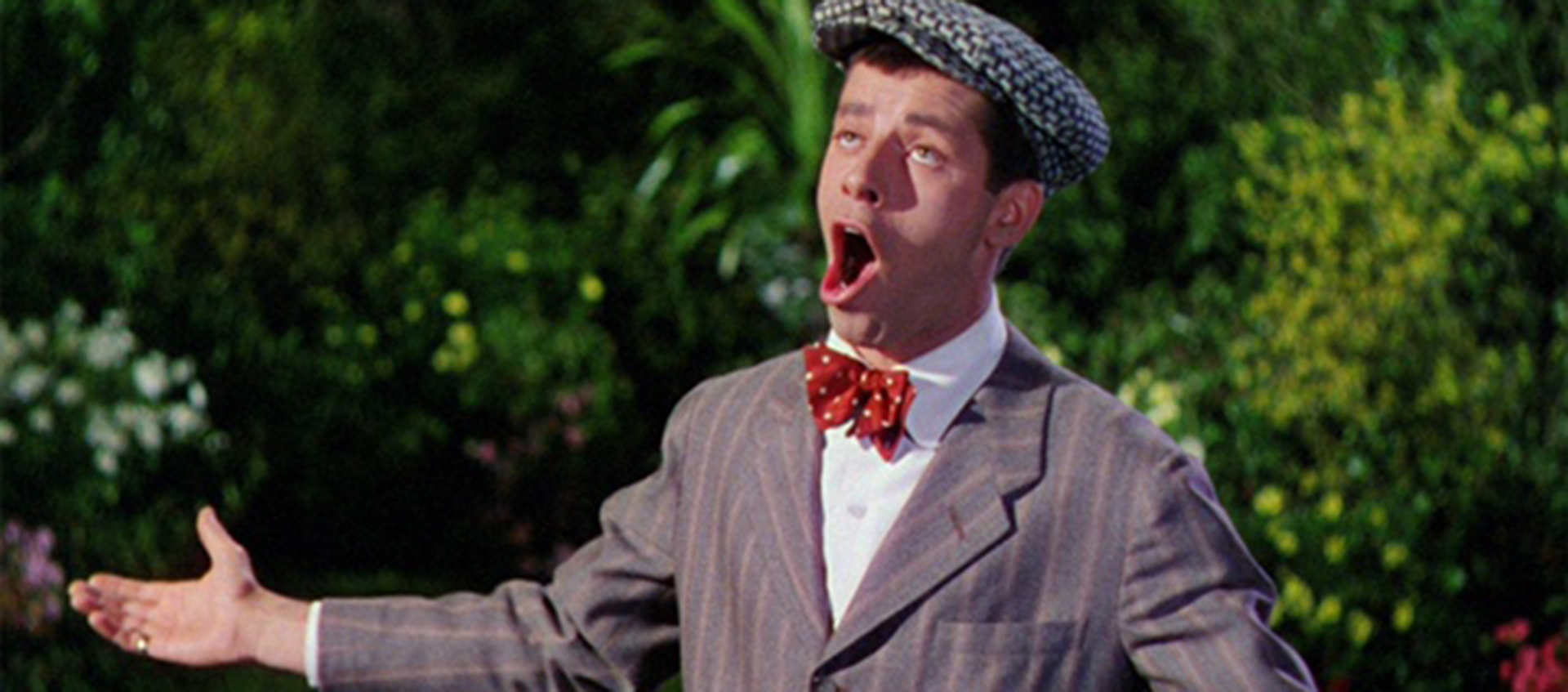  A white man wearing a suit, bowtie, and a fedora holds out his arms and sings. There is foliage behind him.