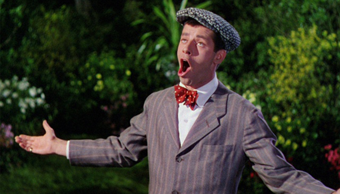  A white man wearing a suit, bowtie, and a fedora holds out his arms and sings. There is foliage behind him.