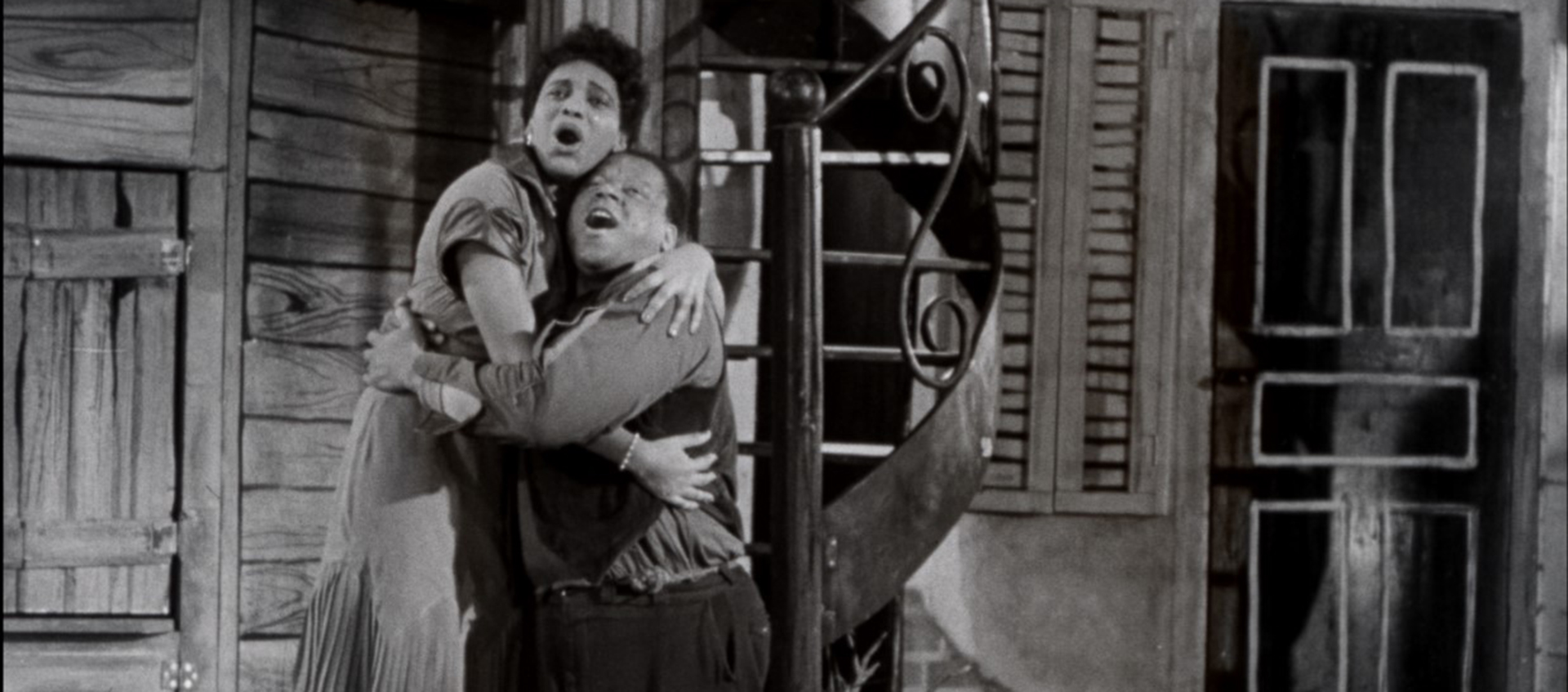 Black-and-white still of a Black woman and a Black man embracing in front of a wooden house.