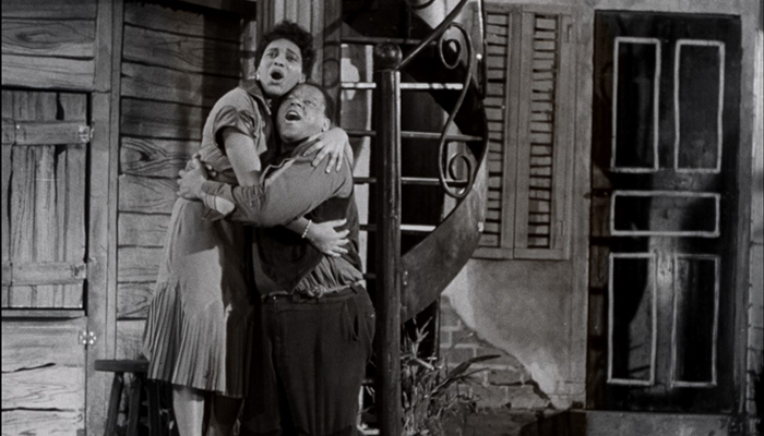 Black-and-white still of a Black woman and a Black man embracing in front of a wooden house.