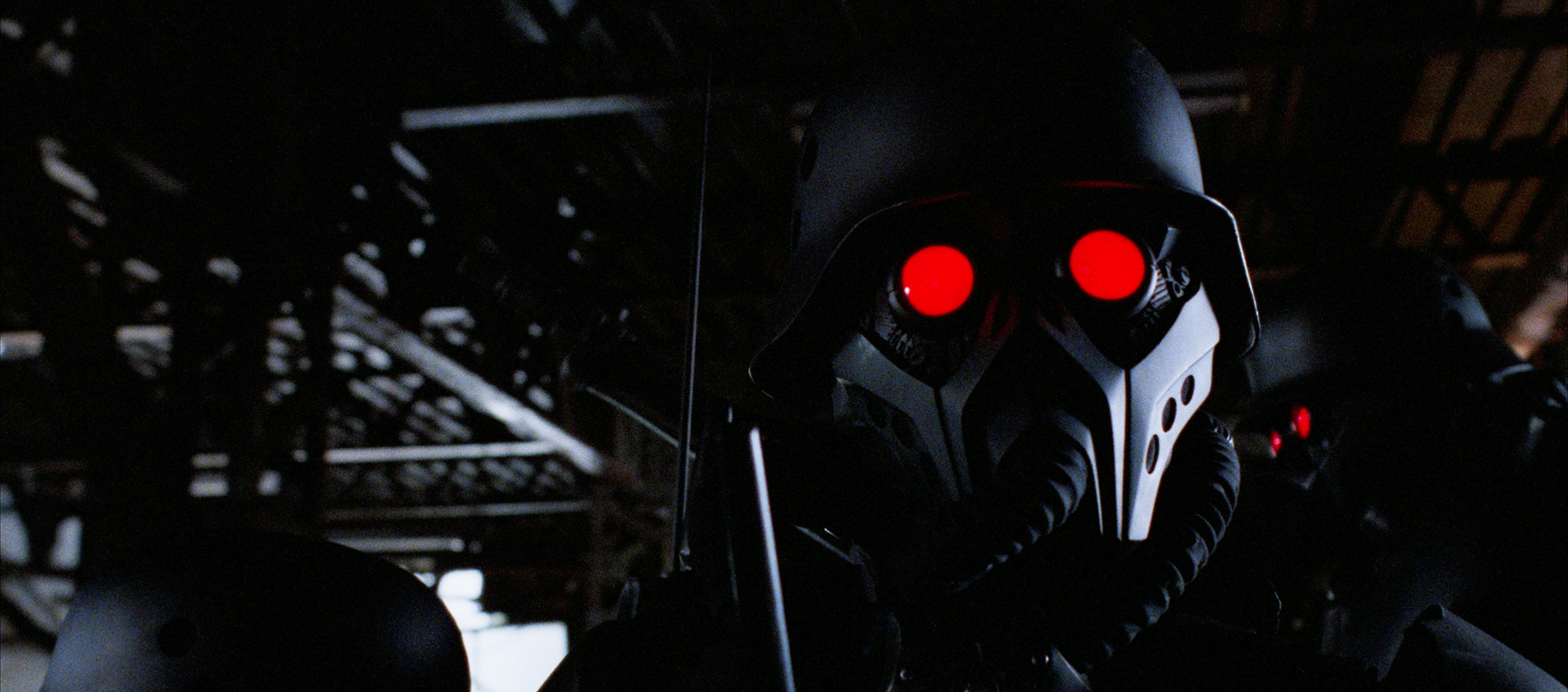 A figure in a black armored suit looks into the camera with bright red eyes.