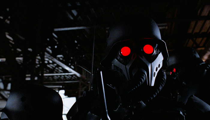 A figure in a black armored suit looks into the camera with bright red eyes.