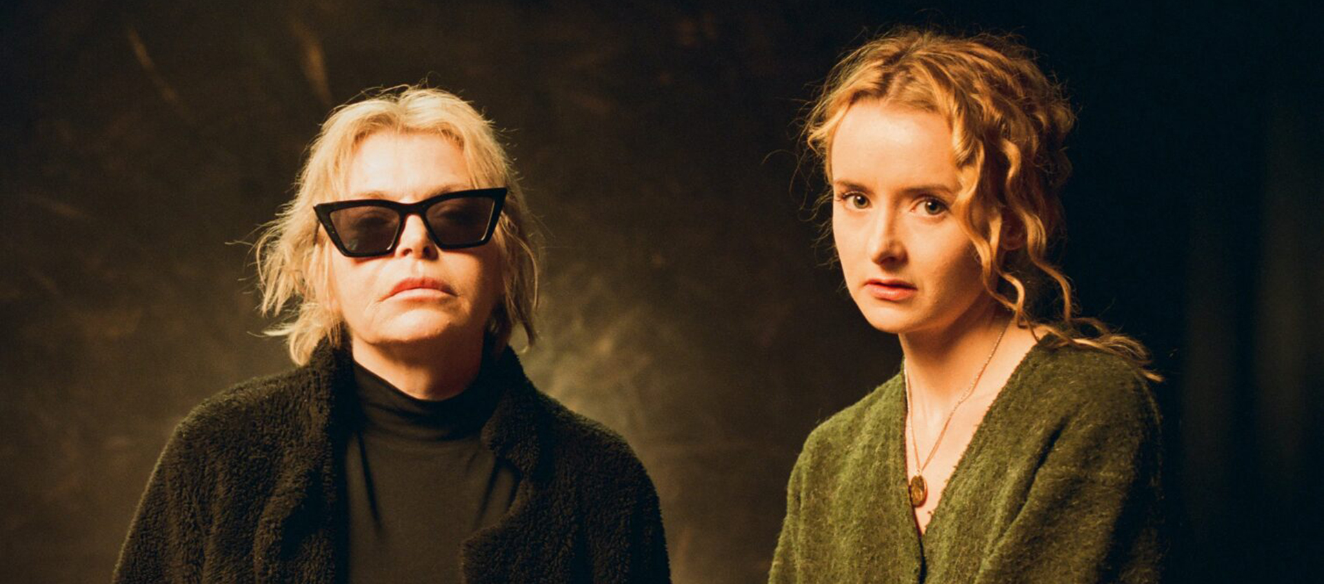 A still of two blonde white women. The woman of the right wears glasses and is older than the woman on the right.