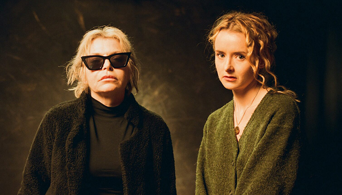 A still of two blonde white women. The woman of the right wears glasses and is older than the woman on the right.