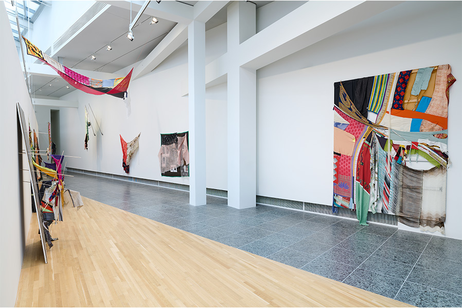 A gallery space featuring an installation of colorful fabric sculptures and paintings hanging from the walls and ceiling.