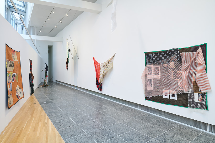 A gallery space featuring an installation of colorful fabric paintings hanging from the walls.